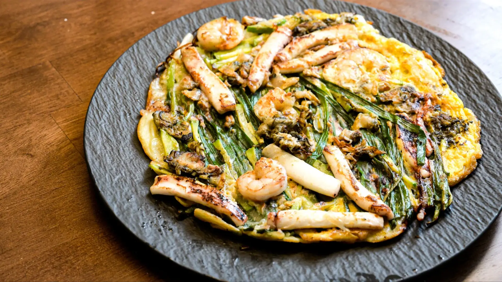 Golden crispy Korean haemul pajeon seafood scallion pancake served on slate plate with squid and scallions.