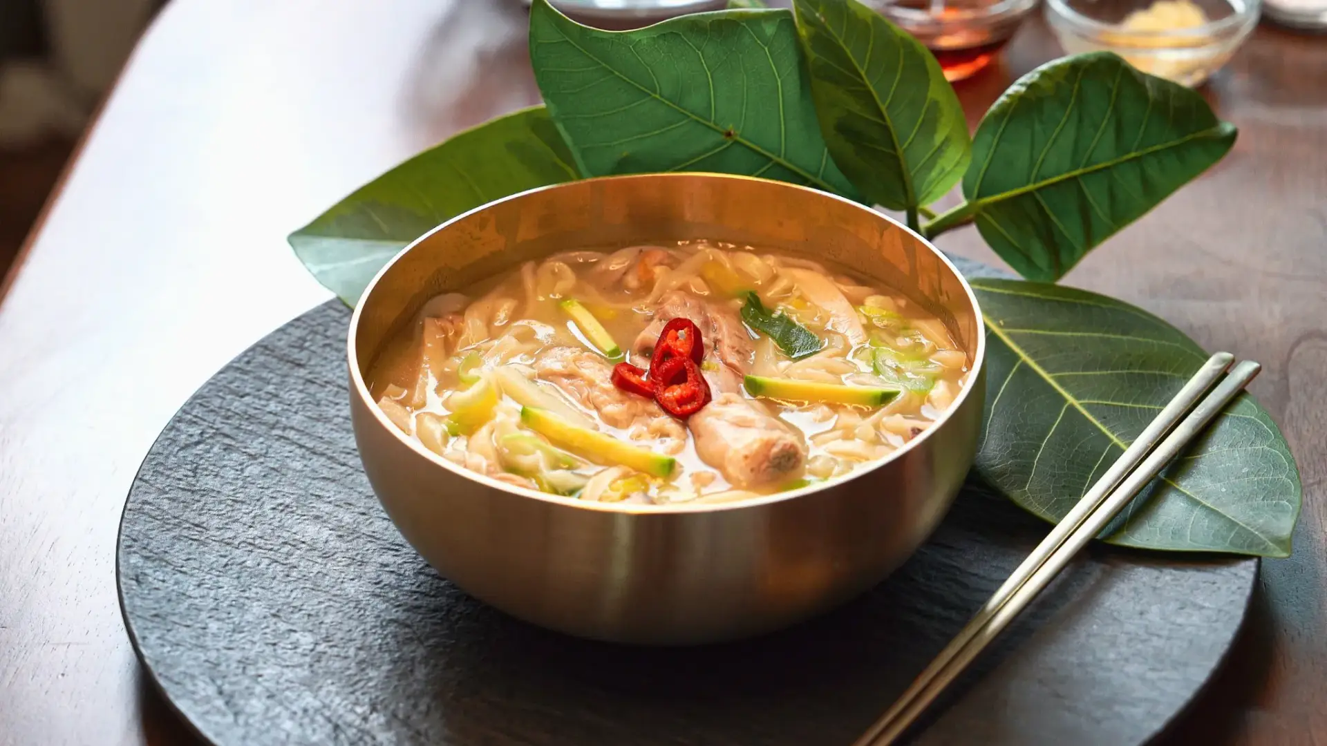 Traditional Korean dak kalguksu chicken noodle soup served in traditional korean bronzeware bowl with knife-cut noodles, tender chicken pieces, and fresh green onion garnish.