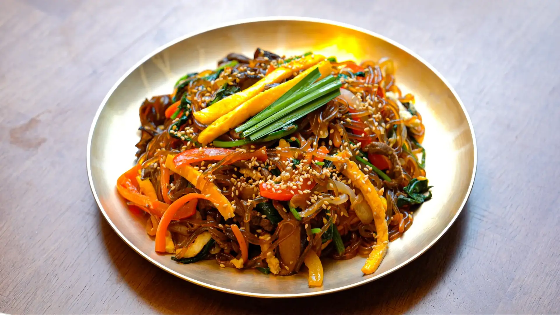 Authentic Korean japchae glass noodles stir-fried with colorful vegetables including carrots and spinach, served on a traditional Korean banjja plate as a traditional Korean dish.