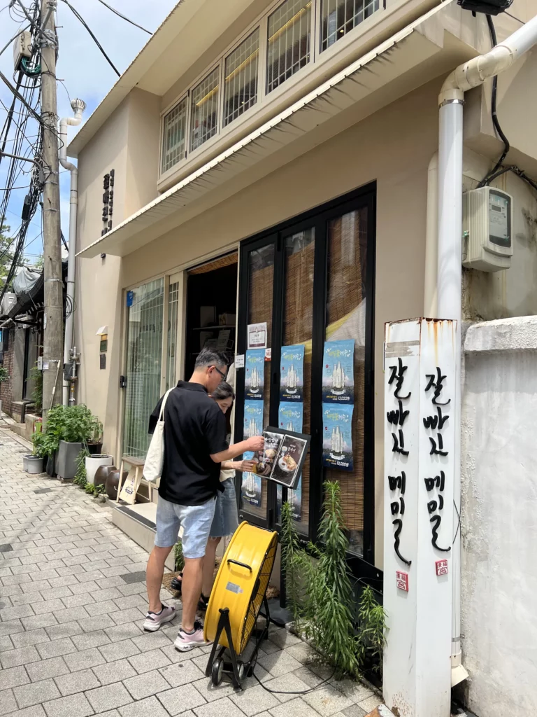 Narrow Seoul alley street view leading to Jalppaejin Memil traditional Korean buckwheat noodle restaurant with outdoor seating and typical urban neighborhood atmosphere.