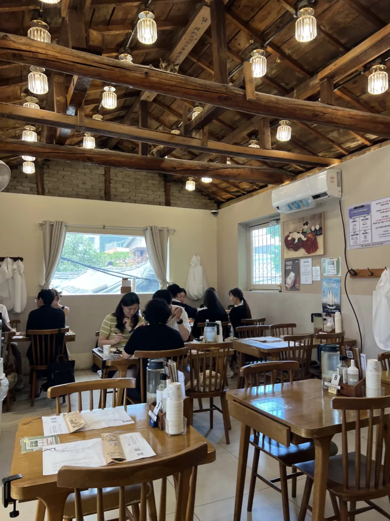 Traditional Korean restaurant interior at Jalppaejin Memil Seoul featuring rustic wooden beam ceiling with pendant lights and diners enjoying handmade buckwheat makguksu noodles.
