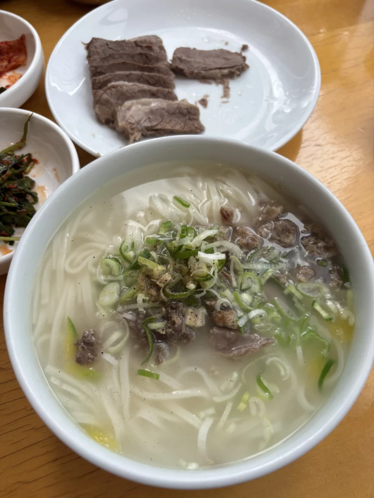 Traditional Korean noodles Andong guksi served in clear beef broth with scallions and hanwoo beef garnish at Sohojeong Seoul restaurant with boiled beef suyuk in background.