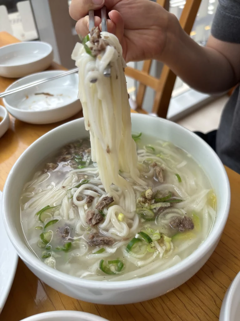 Hand lifting traditional Andong guksi Korean noodles with chopsticks from clear hanwoo beef broth at Sohojeong presidential legacy restaurant in Seoul.