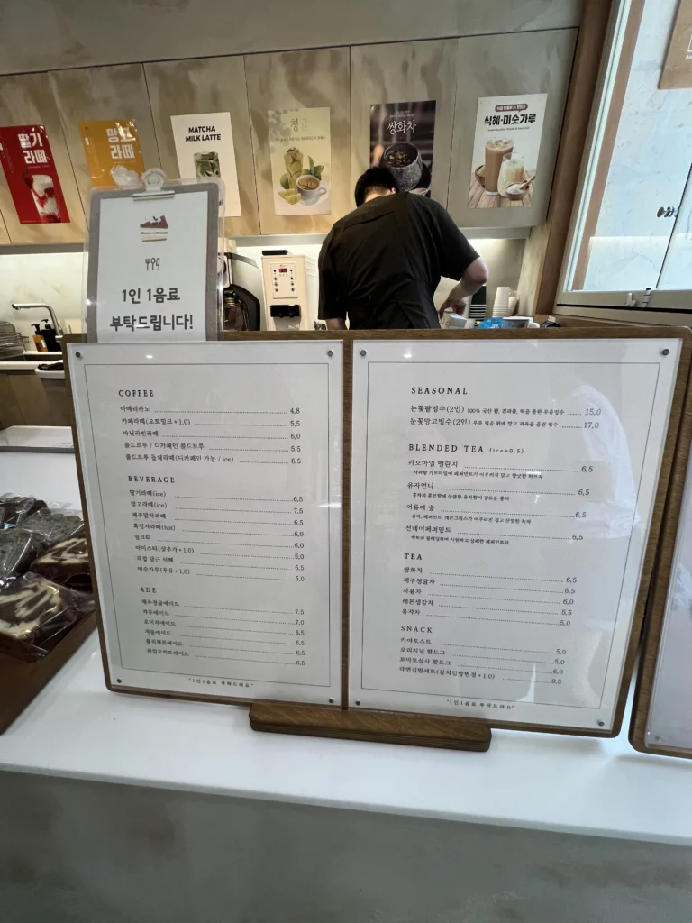 Menu board at Bukhansan Lounge cafe displaying coffee, beverages, seasonal drinks, blended tea, and tea options with barista preparing drinks at the counter in Seoul mountain view cafe.