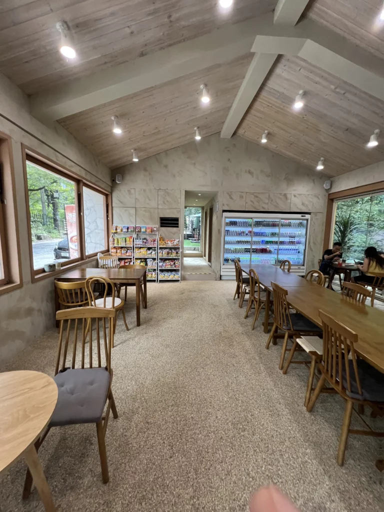 Interior view of Bukhansan Lounge cafe showing vaulted ceiling with pendant lights, wooden tables and chairs, beverage refrigerator, and large windows on both sides offering forest views in Ui-dong Seoul.