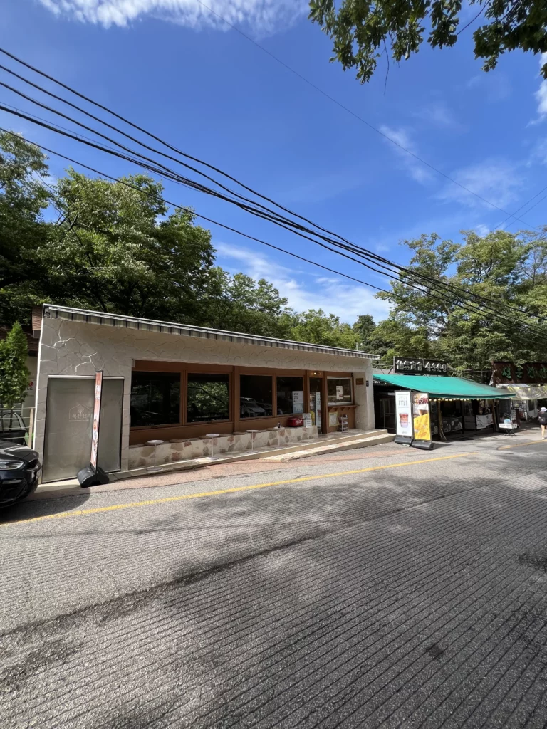 Exterior facade of Bukhansan Lounge mountain view cafe with stone counter seating and parking area at the entrance of Bukhansan National Park in Ui-dong Seoul on a sunny day.