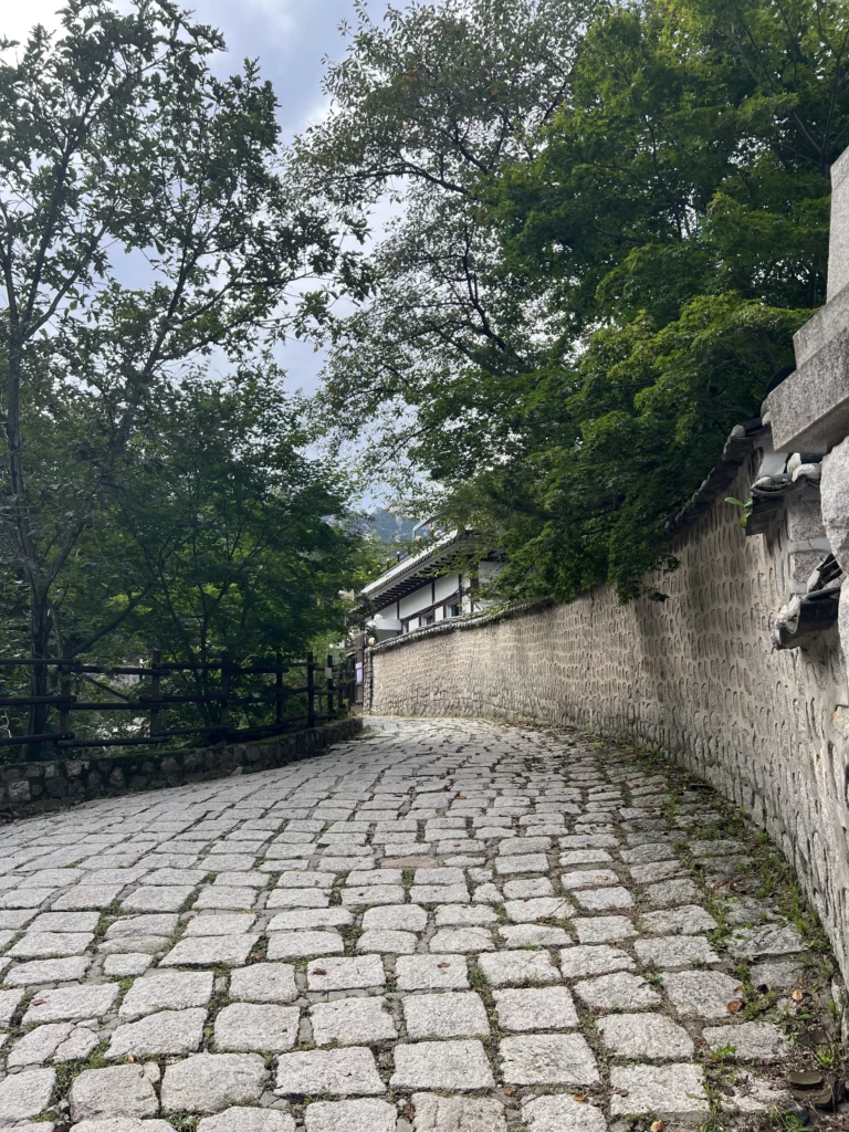 Traditional cobblestone pathway along stone walls leading to Seonungak hanok cafe with Bukhansan Mountain forest backdrop in Seoul.