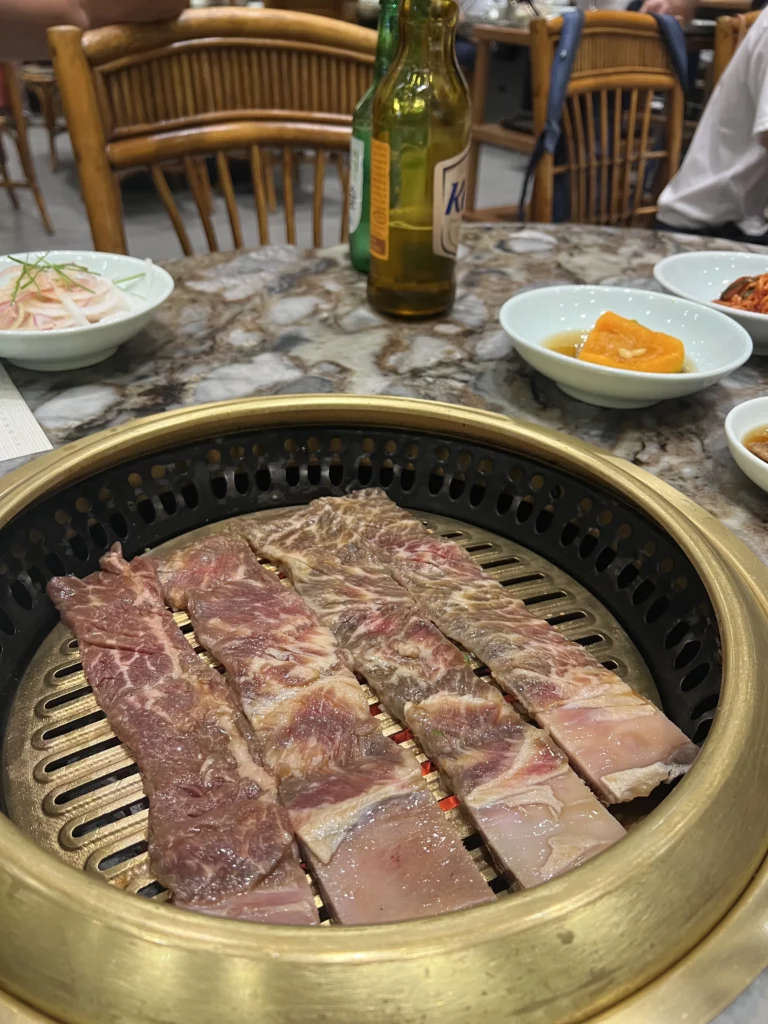Hanwoo galbi beef short ribs grilling on a traditional charcoal BBQ grill at Samwon Garden Seoul, surrounded by Korean banchan side dishes including kimchi and pickled radish.