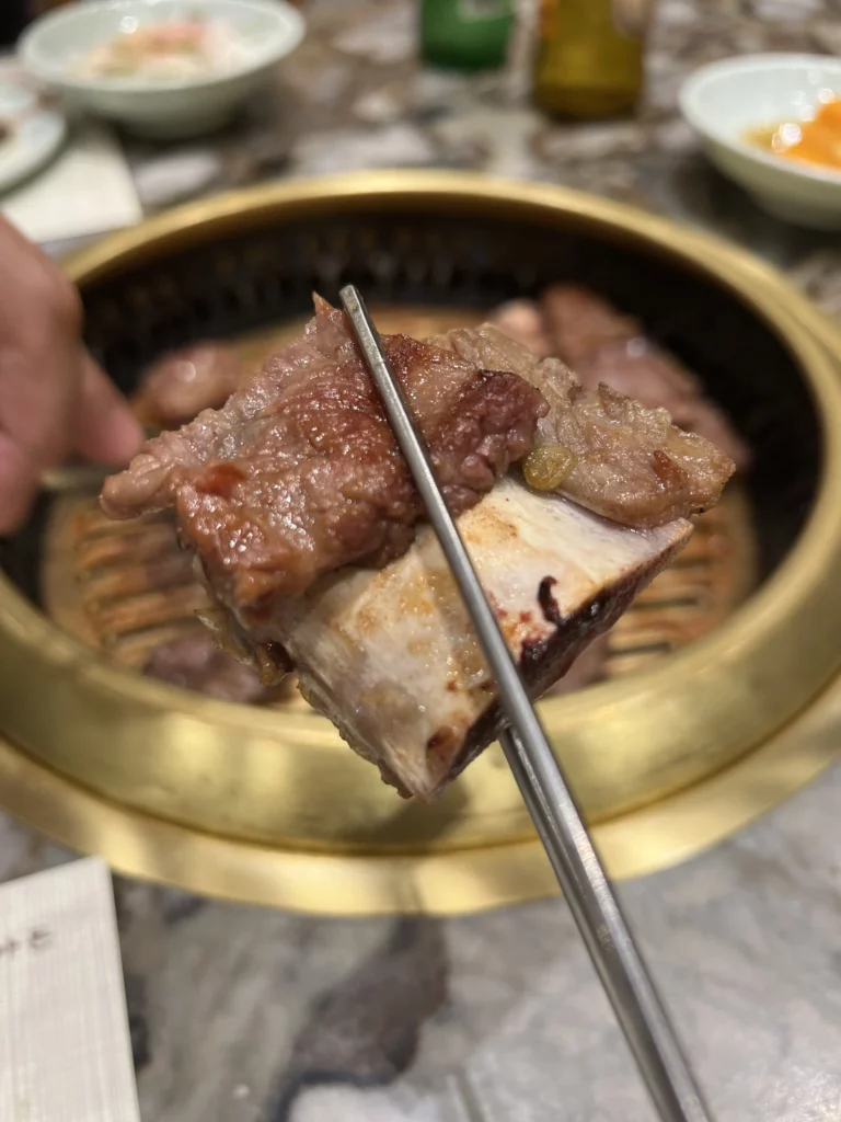 Grilled Hanwoo galbi beef short ribs and vegetables held with chopsticks over charcoal BBQ grill at Samwon Garden Seoul Korean restaurant.
