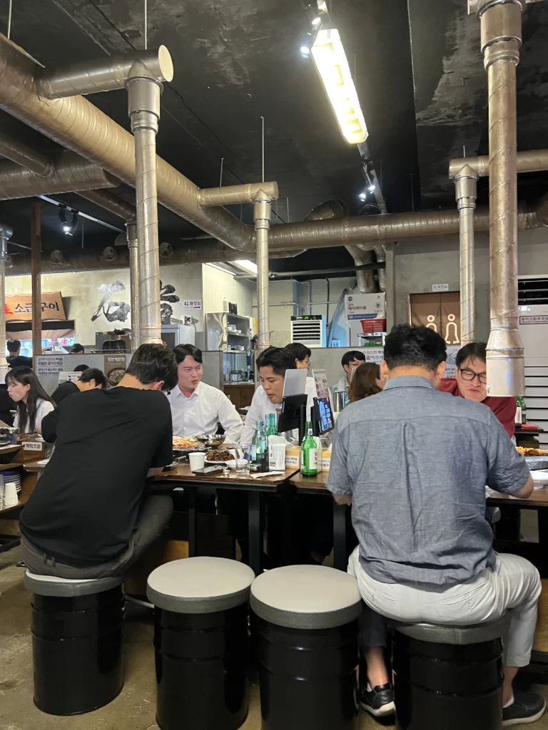 Customers dining at Seokam Saeng Sogeum-gui with industrial-style exposed ductwork and rustic interior design featuring volcanic stone grill tables in Seoul.