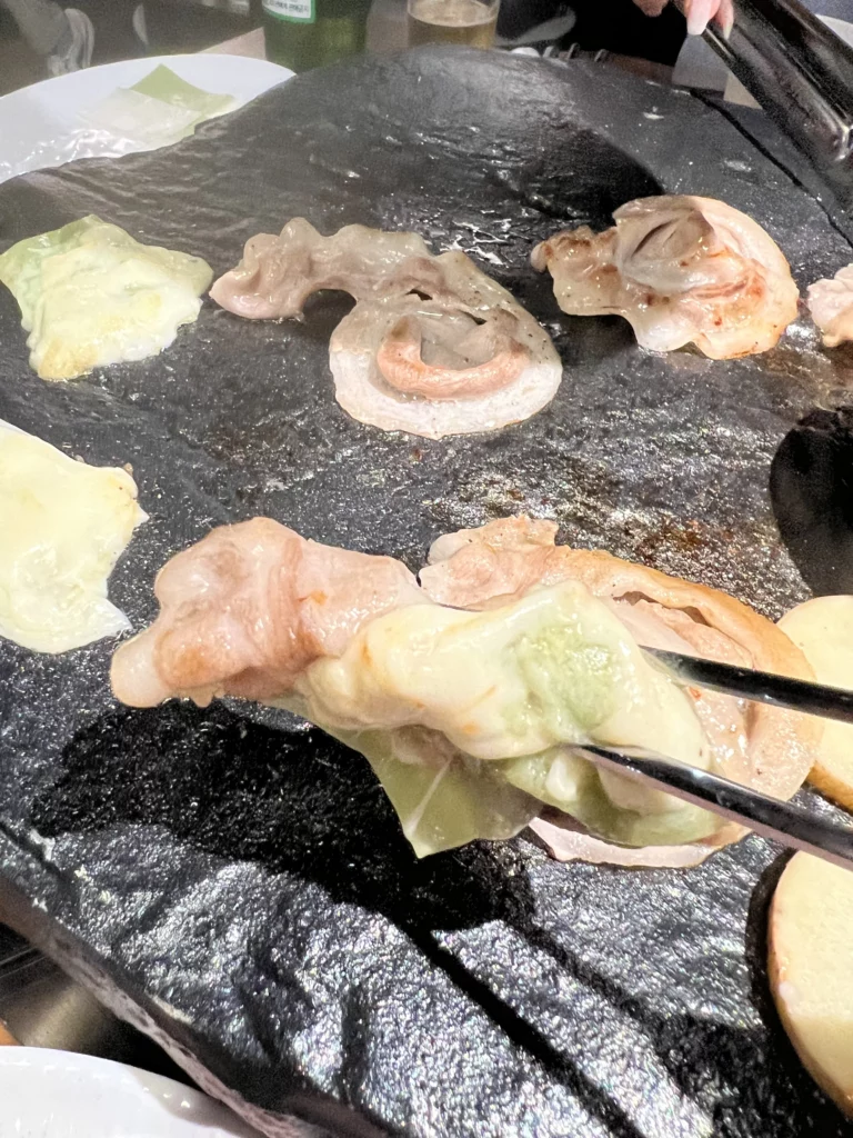 Chopsticks picking up grilled samgyeopsal pork belly with rice cakes from volcanic stone grill plate at Seokam Saeng Sogeum-gui Korean BBQ Seoul.