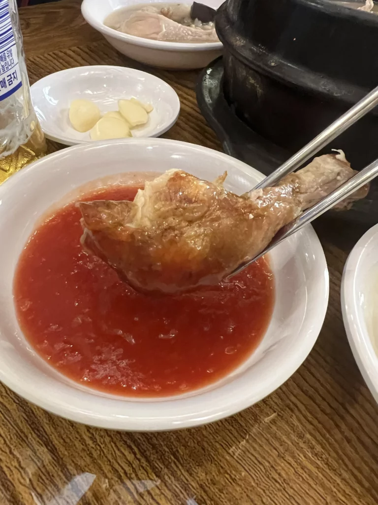 Crispy rotisserie chicken from Tosokchon being dipped in spicy gochujang sauce, traditional Korean eating method at Seoul's legendary samgyetang restaurant near Gyeongbokgung Palace.
