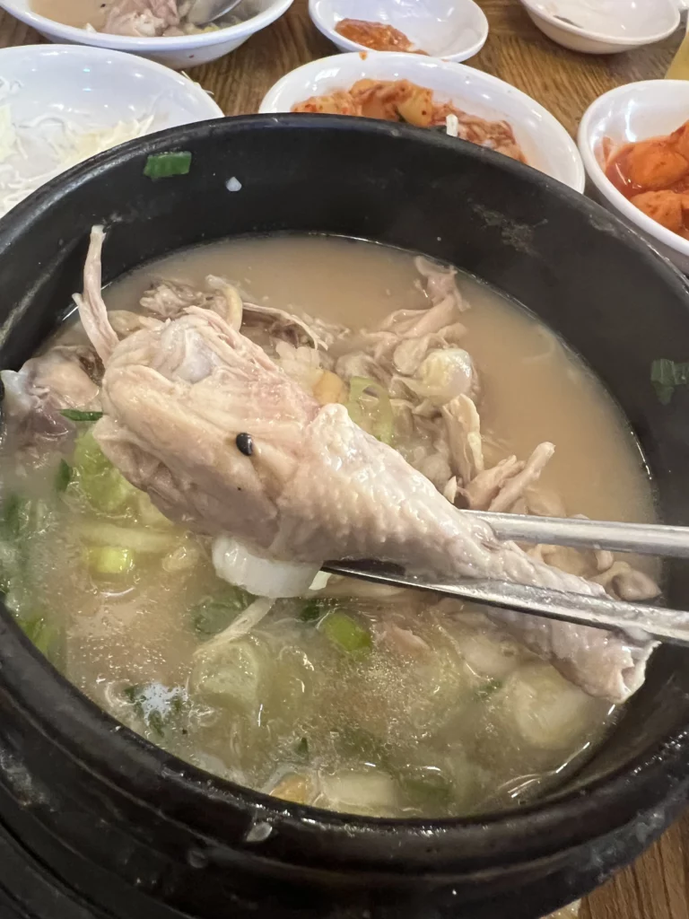 Partially eaten Tosokchon samgyetang in black earthenware pot showing shredded tender chicken in milky ginseng broth with green onions, Seoul's traditional Korean wellness soup.