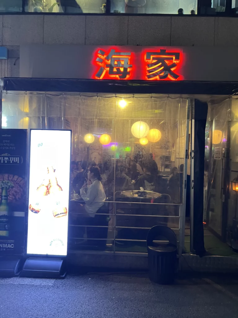 Haega Jjukkumi storefront window view at night showing cozy interior with yellow pendant lights and diners enjoying spicy webfoot octopus jjukkumi at authentic Korean seafood restaurant in Sinsa-dong Gangnam Seoul.