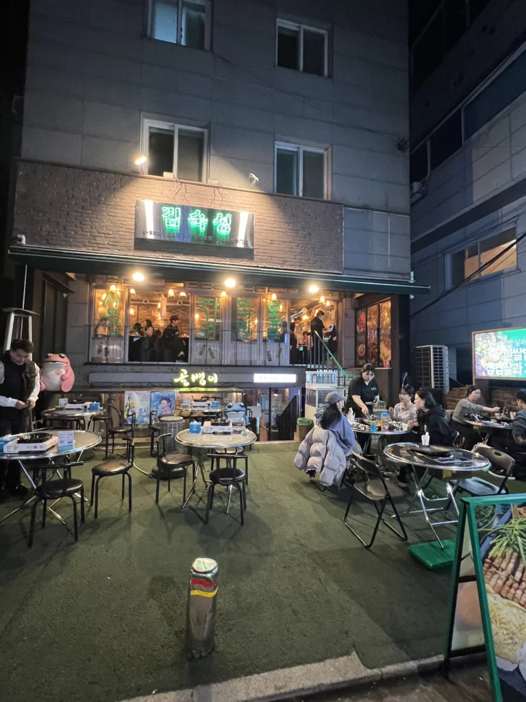 Outdoor seating area at Gimsukseoung restaurant in Sinsa-dong, Seoul, serving 22-day aged samgyeopsal Korean BBQ with dining guests at night.
