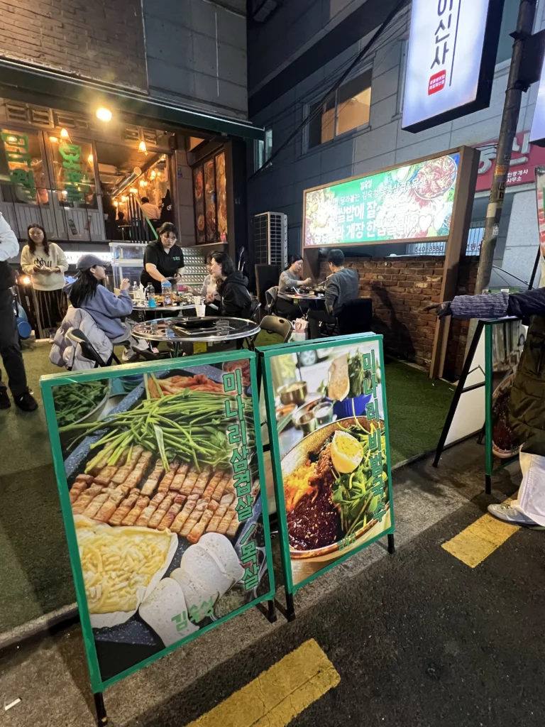 Outdoor menu board displaying 22-day aged samgyeopsal dishes at Gimsukseoung restaurant in Seoul's Sinsa-dong neighborhood with dining area in background.