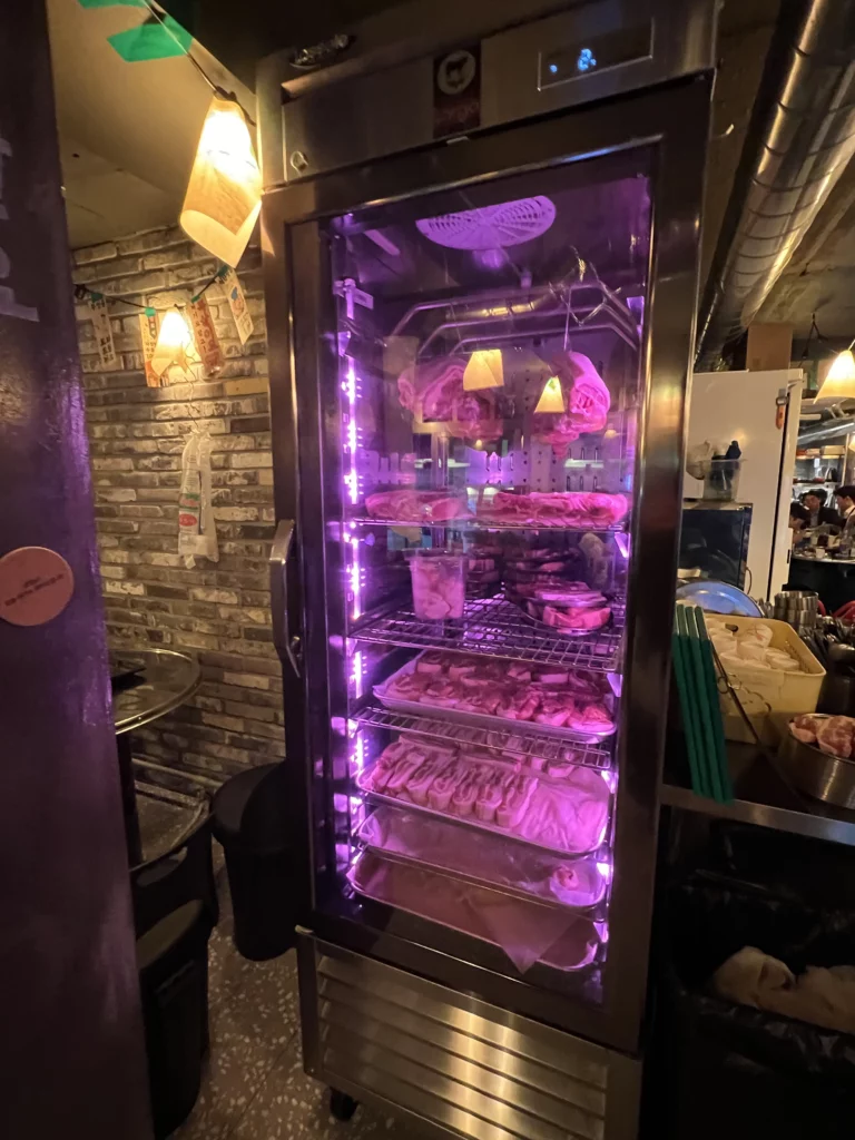 Refrigerated display case with purple LED lighting showcasing 22-day aged samgyeopsal and moksal cuts at Gimsukseoung Korean BBQ restaurant in Seoul.