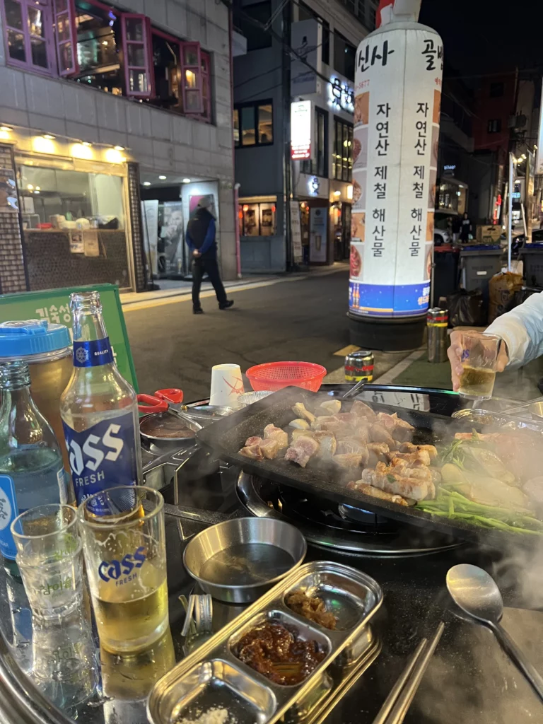 Outdoor Korean BBQ dining at Gimsukseoung in Sinsa-dong, Seoul, featuring 22-day aged samgyeopsal grilling with Cass Fresh beer on a vibrant night street.