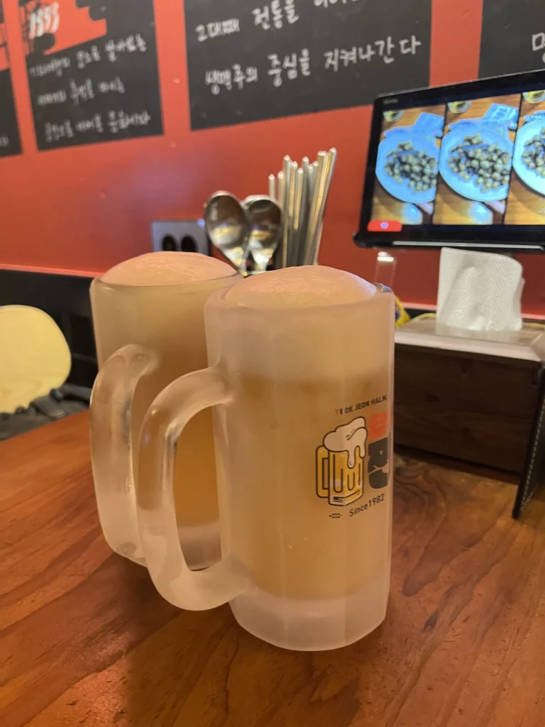 Frosted glass mugs filled with Korean sal-eoreum iced beer at Yeokjeon Halmae Maekju restaurant with traditional Korean menu board in background.