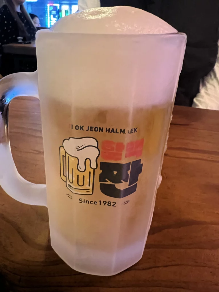 Close-up of overflowing Korean sal-eoreum iced beer in branded frosted mug at Yeokjeon Halmae Maekju establishment since 1982.