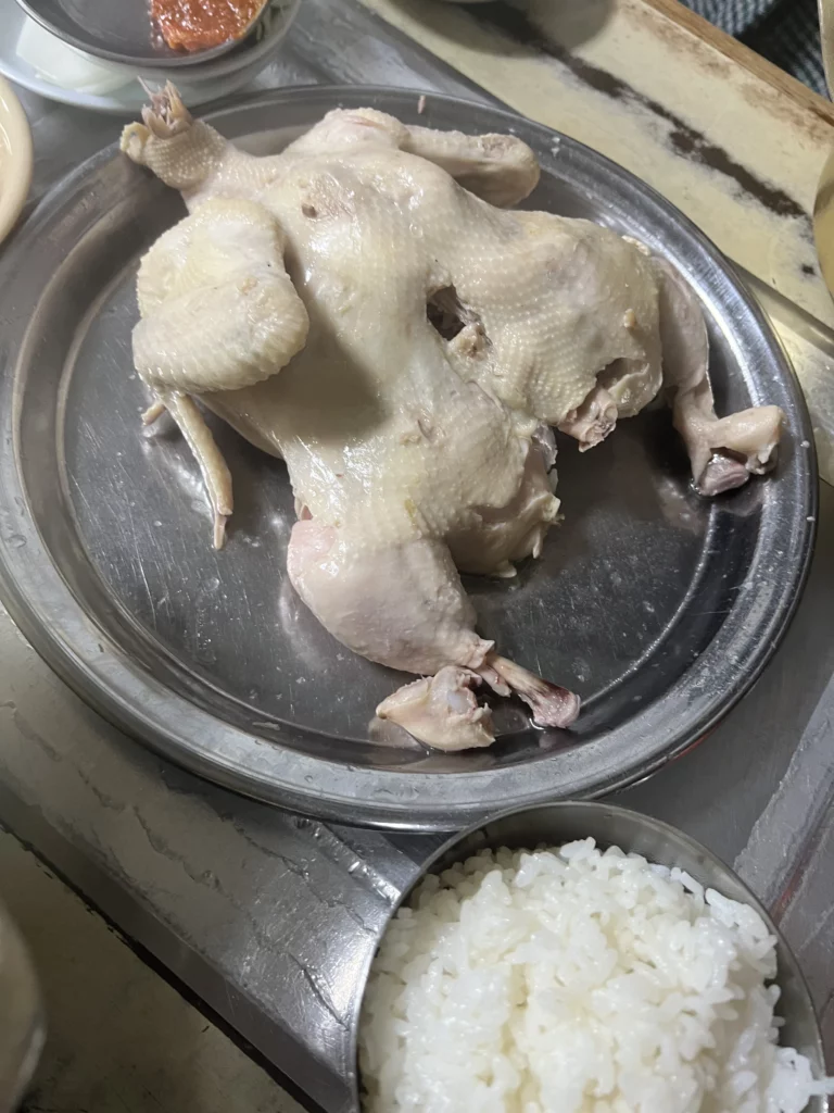 Whole dak baeksuk chicken served on metal plate at Sarangbang Kalguksu restaurant in Seoul with garlic cloves in traditional Korean boiled chicken soup preparation.