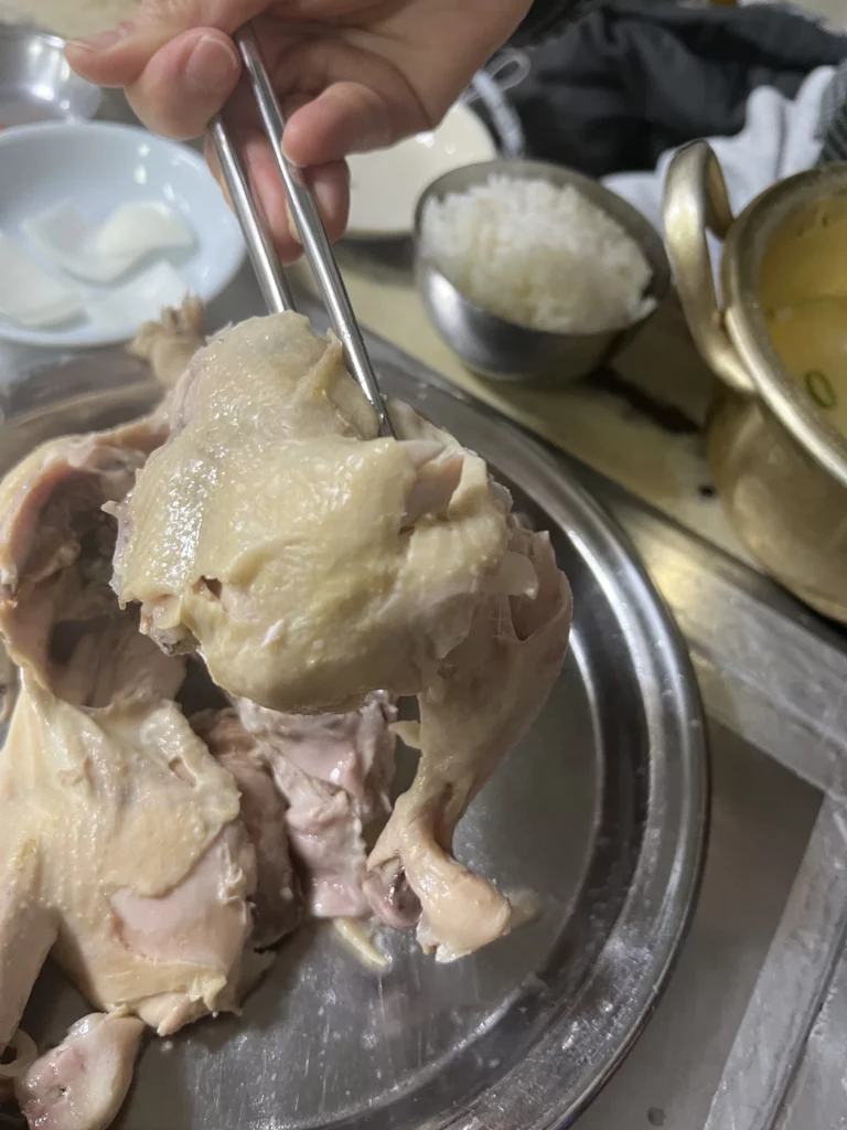 Tender chicken piece held with chopsticks from dak baeksuk at Sarangbang Kalguksu demonstrating traditional Korean eating method for boiled chicken soup.
