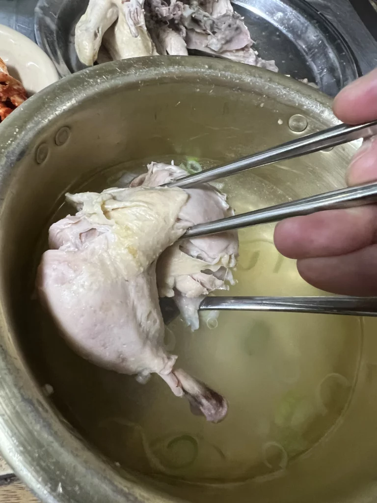 Tender boiled chicken being lifted with chopsticks from garlic-infused dak baeksuk broth at Sarangbang Kalguksu demonstrating traditional Korean chicken soup eating method in Seoul.