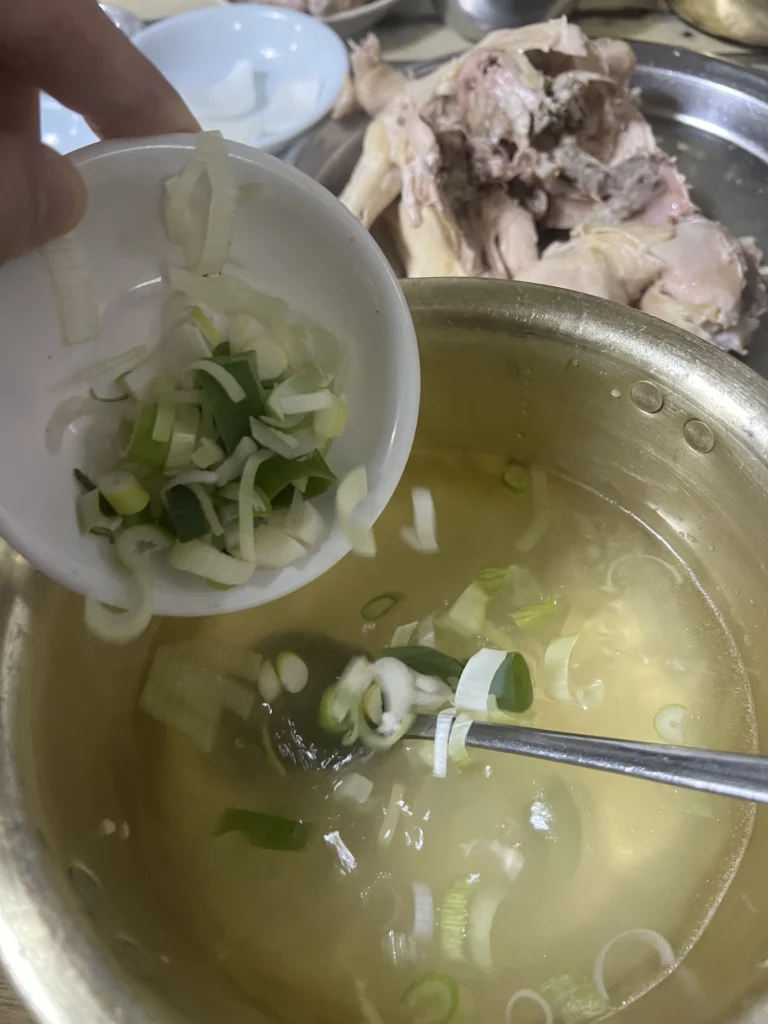 Adding green onions to garlic-infused dak baeksuk broth with torn chicken at Sarangbang Kalguksu traditional Korean boiled chicken soup restaurant in Seoul.