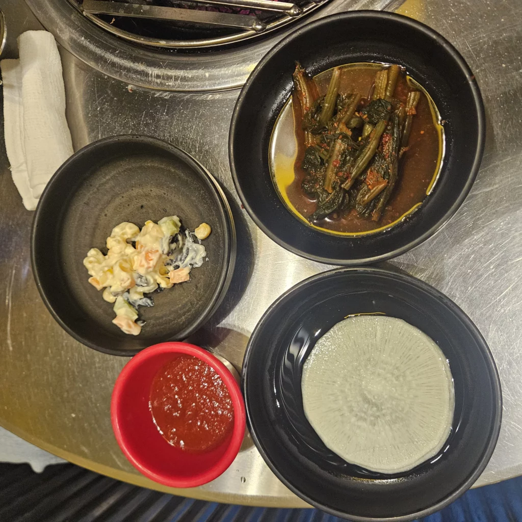 Assorted Korean side dishes served with short ribs, including pickled radish slices, marinated greens, macaroni salad, and a red dipping sauce in small bowls on a metal restaurant table.