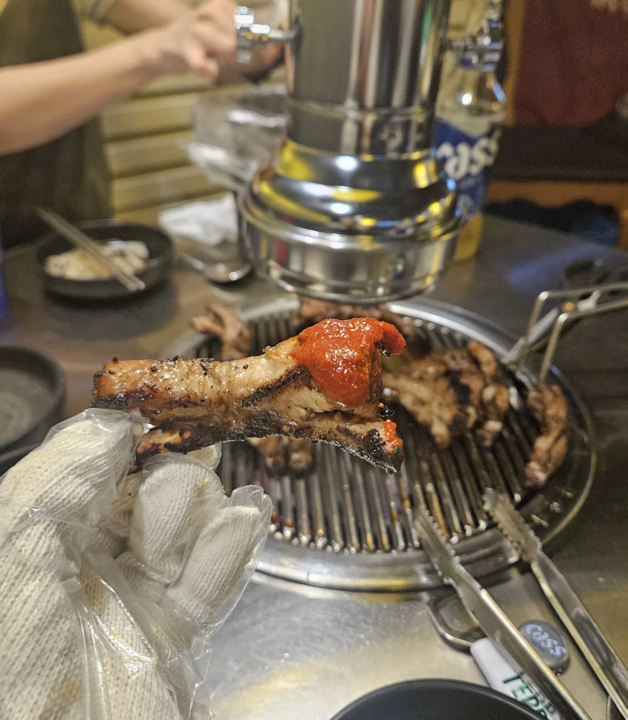 Grilled short ribs held with a gloved hand, topped with a red sauce, with more meat cooking on a metal Korean BBQ grill in the background, tongs and side dishes visible on the table.