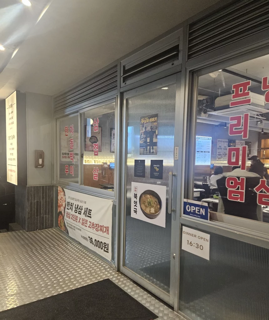 Frozen Samgyeopsal Korean BBQ restaurant entrance with glass doors, menu posters, and banners promoting meal sets, offering a clear view into the warmly lit dining area inside.