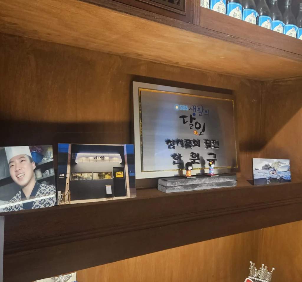 A warm wooden display inside a Frozen Samgyeopsal Korean BBQ restaurant, showing framed photos, an SBS award plaque, rows of soju bottles, and lucky cat figurines arranged neatly on the shelves, highlighting the restaurant’s cozy and character-filled interior décor.