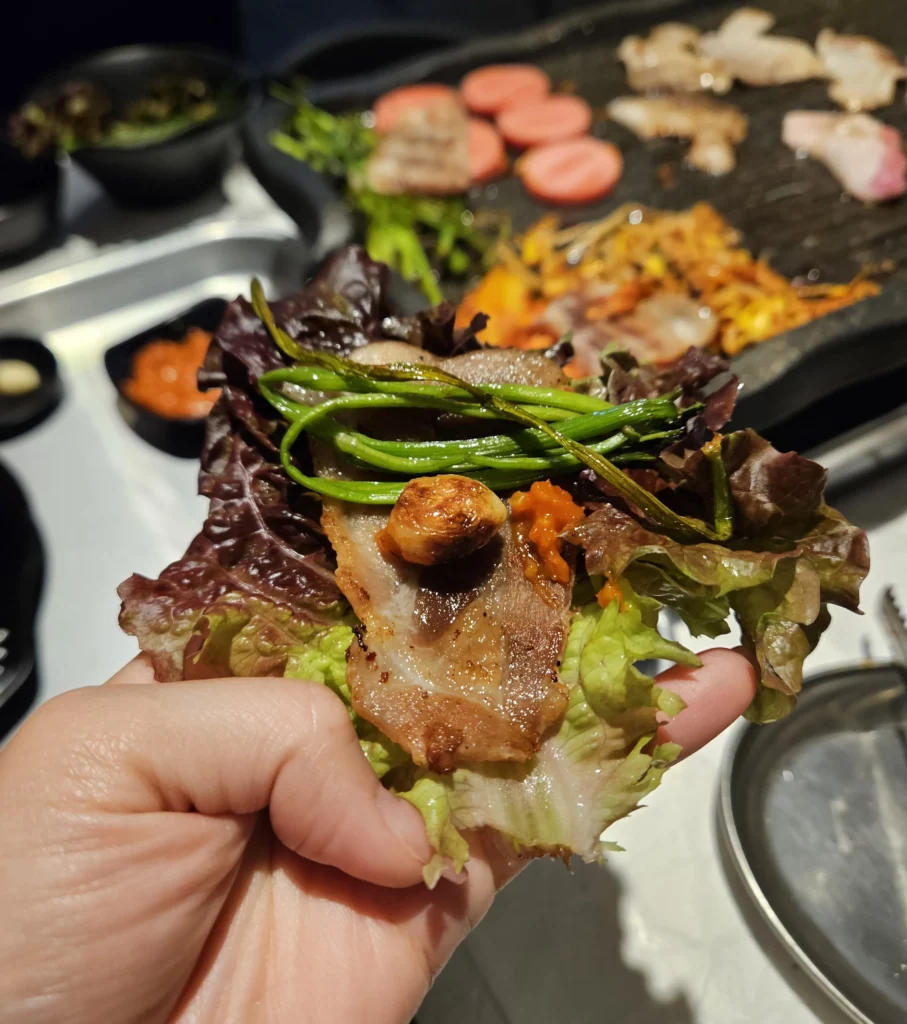 A hand holding a ssam wrap at a Frozen Samgyeopsal Korean BBQ restaurant, filled with grilled pork belly, sautéed greens, roasted garlic, and spicy ssamjang inside a fresh lettuce leaf, with the grill and side dishes visible in the background.
