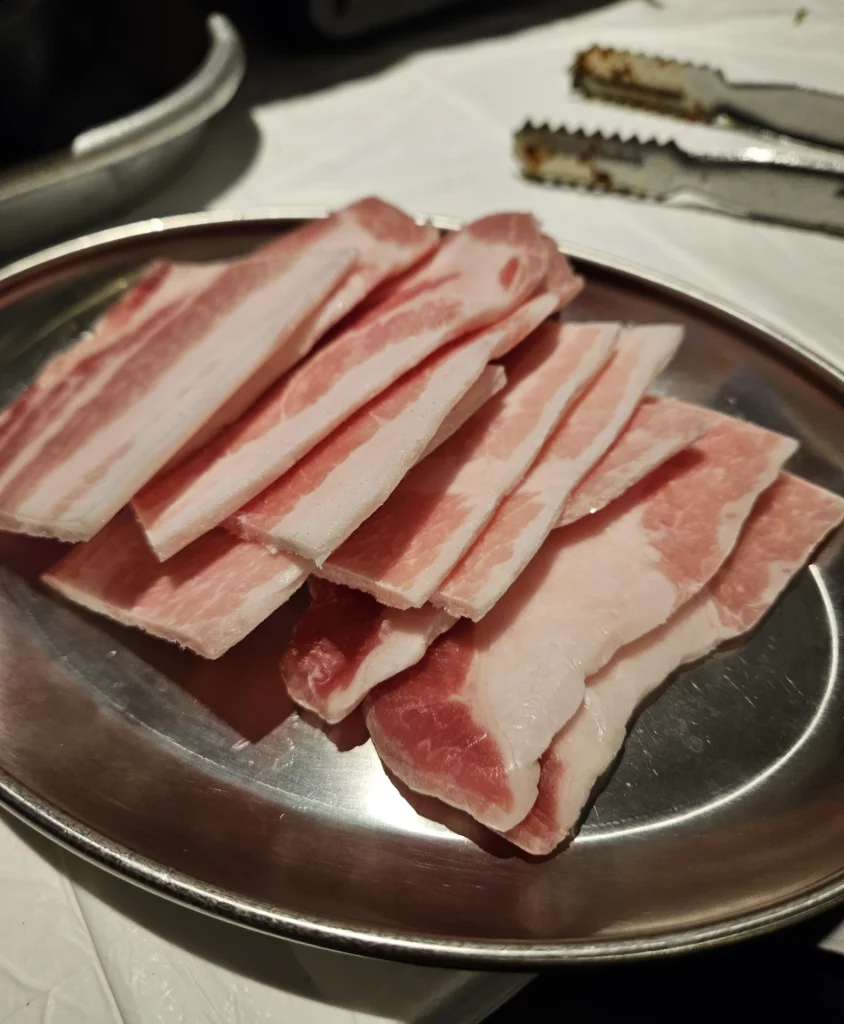 A metal tray filled with neatly stacked slices of frozen pork belly for Frozen Samgyeopsal Korean BBQ, showing the pink marbling and thinly cut layers before grilling.