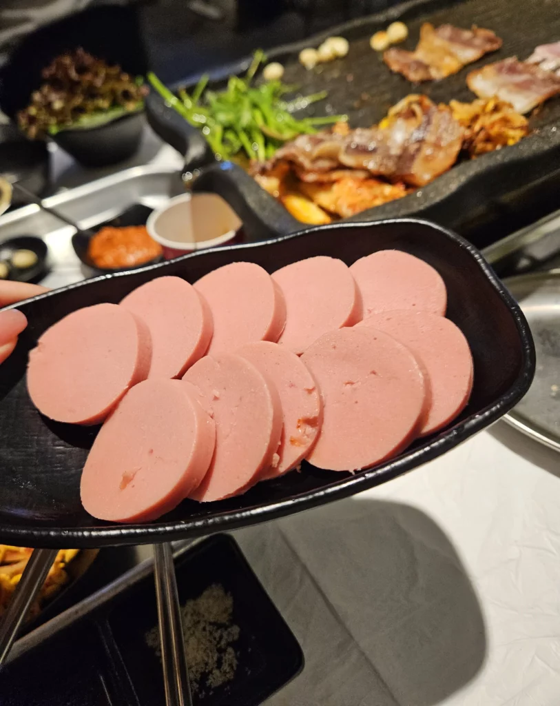 A black plate filled with round sausage slices served as a side dish at a Frozen Samgyeopsal Korean BBQ restaurant, with the grill and sizzling pork belly in the background.