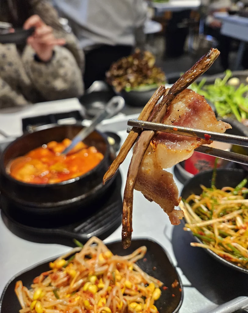 Chopsticks holding a bite of grilled pork belly with stir-fried greens at a Frozen Samgyeopsal Korean BBQ restaurant, with side dishes like bean sprouts and bubbling stew visible in the background.