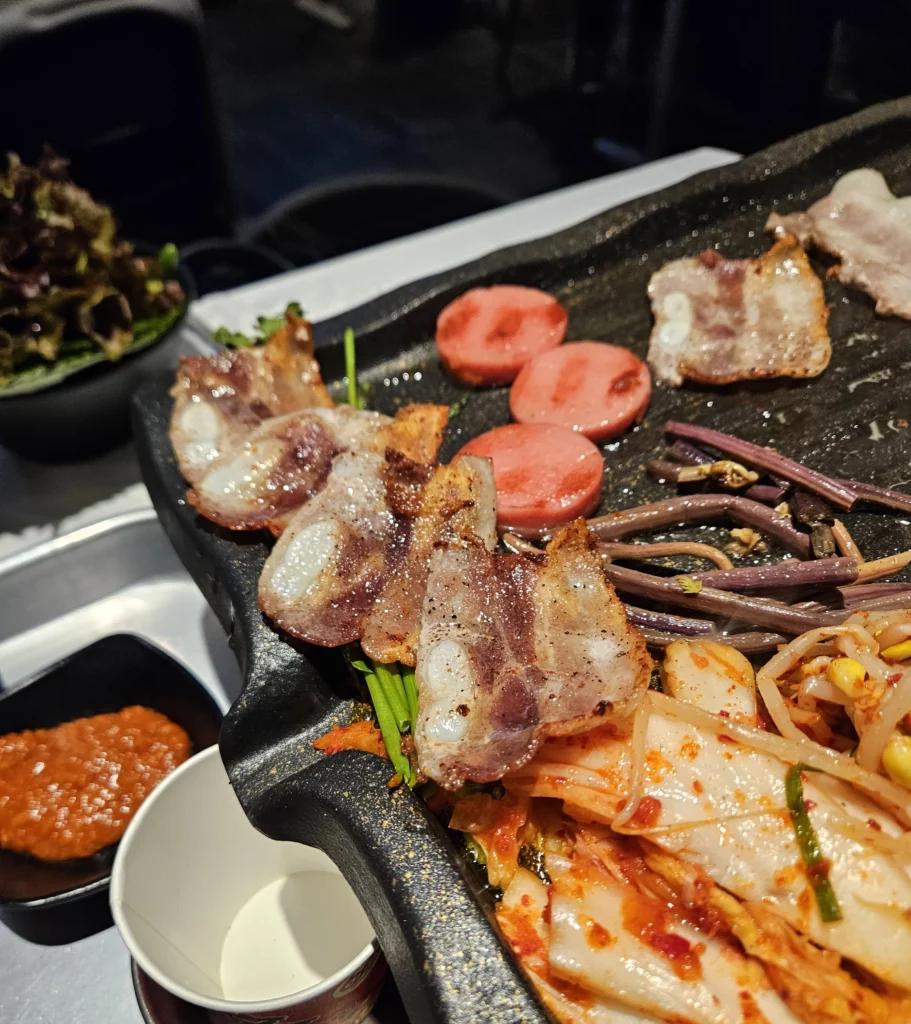 Frozen Samgyeopsal Korean BBQ slices grilled to a golden crisp on a hot plate alongside kimchi, bean sprouts, sausage rounds, and seasoned greens, creating a sizzling and flavorful mix ready to eat.