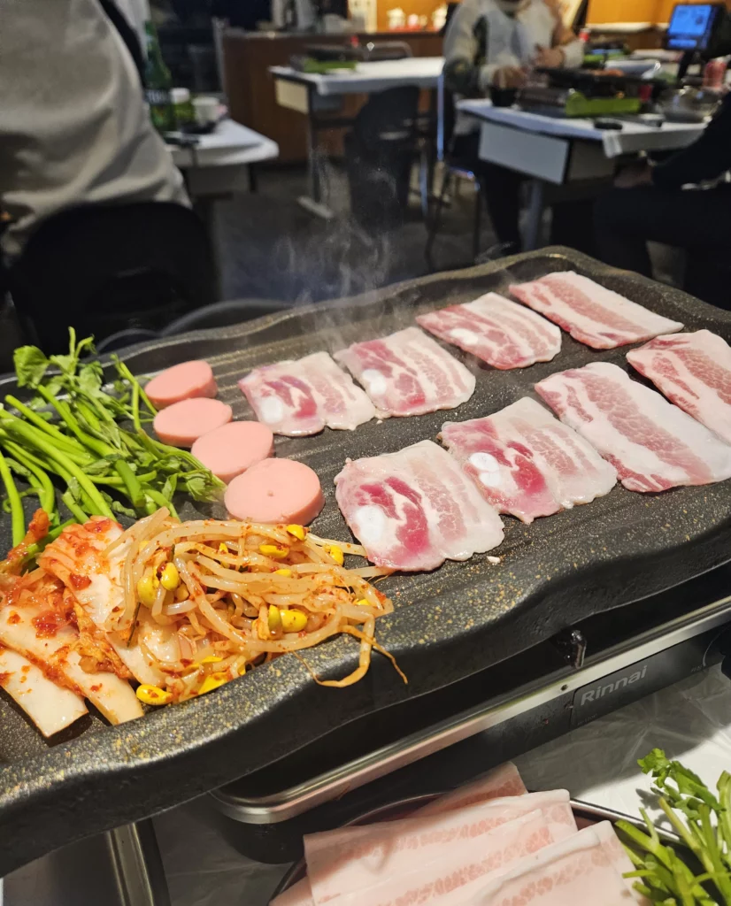 Frozen Samgyeopsal Korean BBQ slices sizzling on a hot grill alongside kimchi, seasoned bean sprouts, sausage slices, and fresh greens, with steam rising as diners cook their meal in a lively restaurant setting.