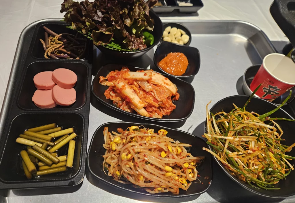 A tray of colorful side dishes served with Frozen Samgyeopsal Korean BBQ, including kimchi, seasoned bean sprouts, spicy green onion salad, pickled garlic stems, sausage slices, fresh lettuce leaves, raw garlic, and ssamjang, arranged neatly in black bowls.