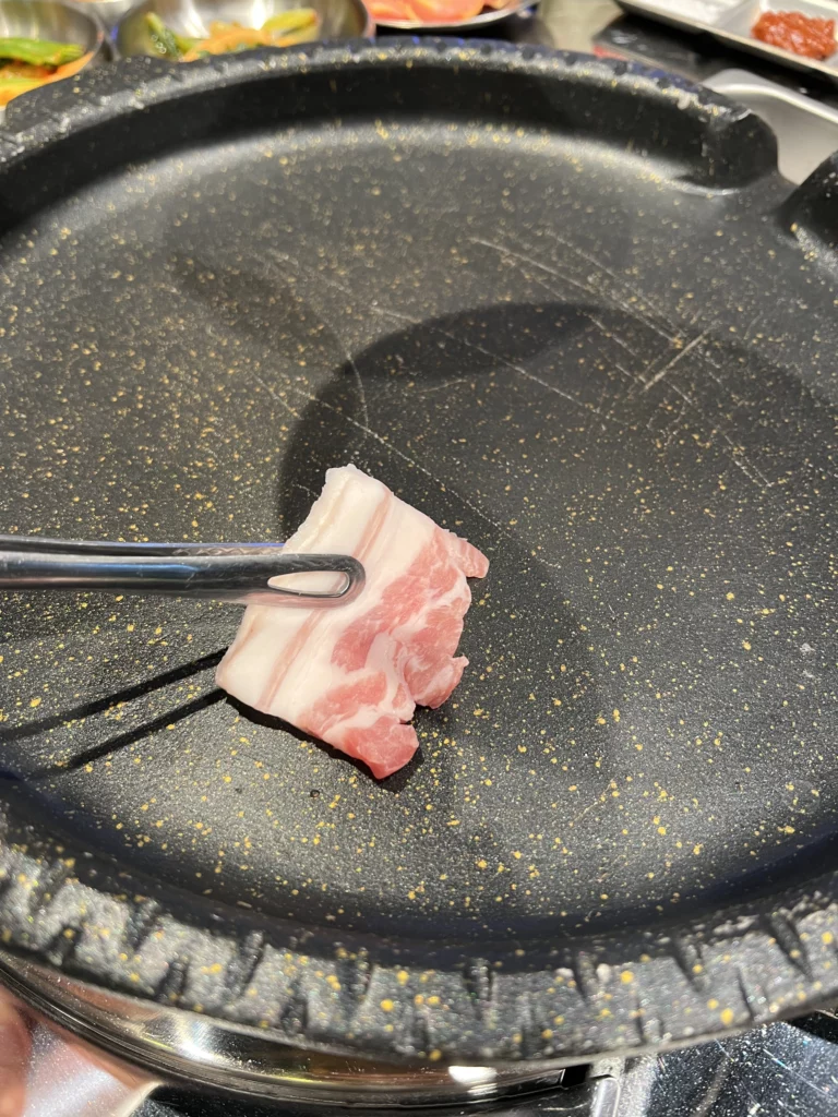 Tongs placing fresh samgyeopsal pork belly slice on hot grill at Moosik authentic Korean BBQ restaurant in Sinsa-dong Gangnam Seoul.