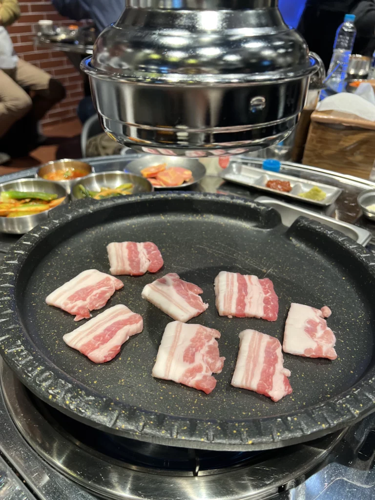 Samgyeopsal pork belly slices grilling on hot plate at Moosik Korean BBQ restaurant with traditional side dishes in Sinsa-dong Gangnam Seoul.