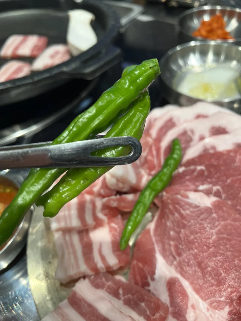 Tongs holding fresh green chili peppers over marbled samgyeopsal pork belly at Moosik Korean BBQ restaurant in Sinsa-dong Gangnam Seoul.
