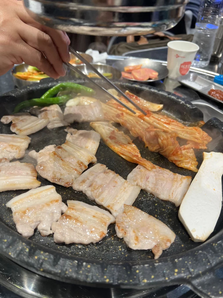 Tongs flipping grilled samgyeopsal pork belly kimchi and vegetables on grill at Moosik authentic Korean BBQ restaurant in Sinsa-dong Gangnam Seoul.