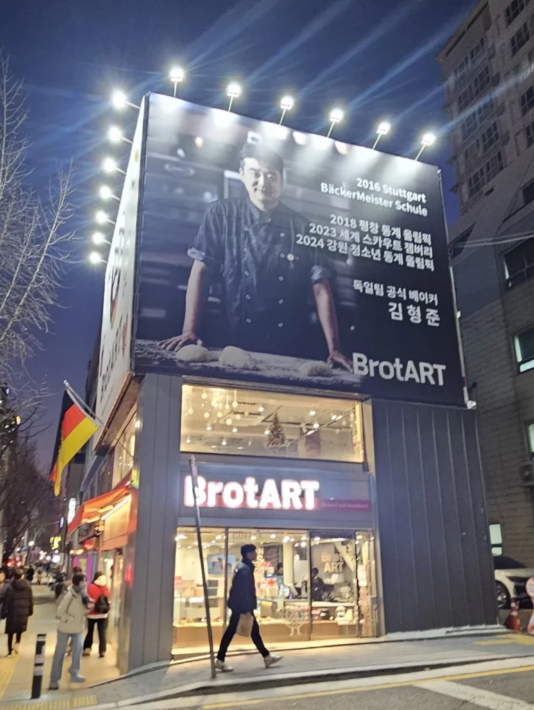 Modern corner bakery cafe exterior at night, featuring a large illuminated billboard, glass storefront, and warm interior lighting in an urban street setting.