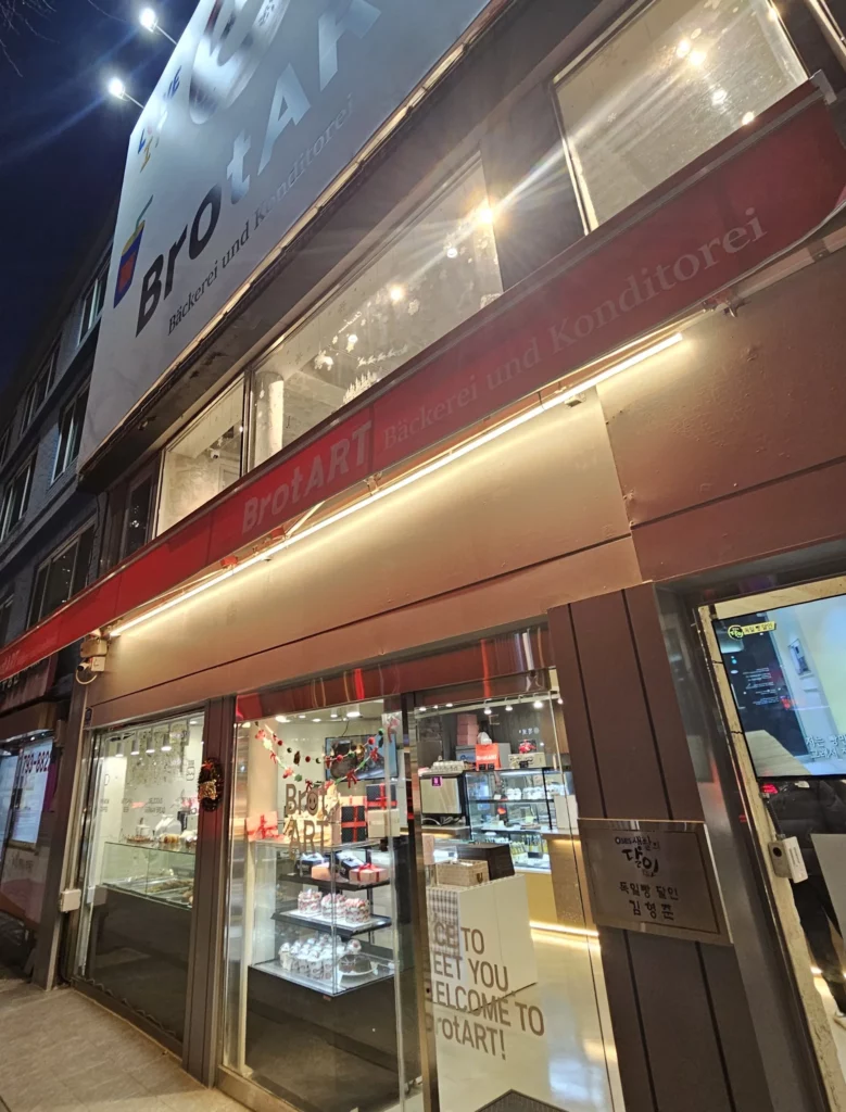 Night exterior of a modern bakery cafe with illuminated signage, large glass windows, and a warm interior showcasing cakes and pastries inside.