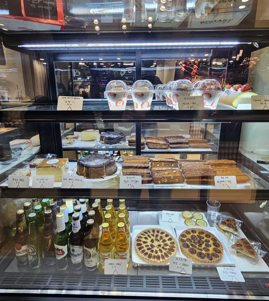 Glass display case inside a bakery cafe featuring assorted cakes, layered desserts, pastries, and bottled drinks neatly arranged on shelves.