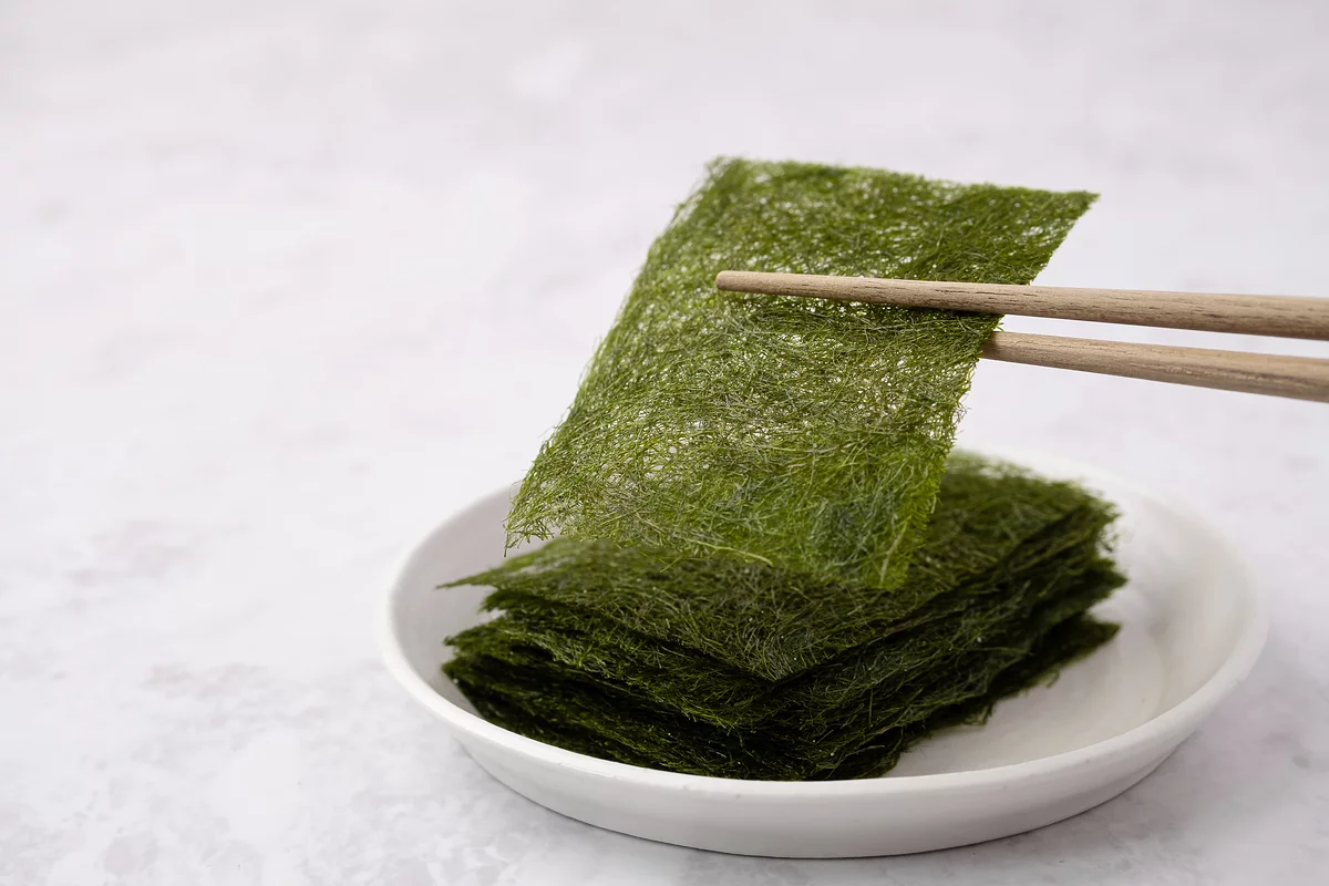 Dried gamtae (Ecklonia cava) Korean seaweed sheets held with chopsticks showing olive-green color and delicate texture on white plate