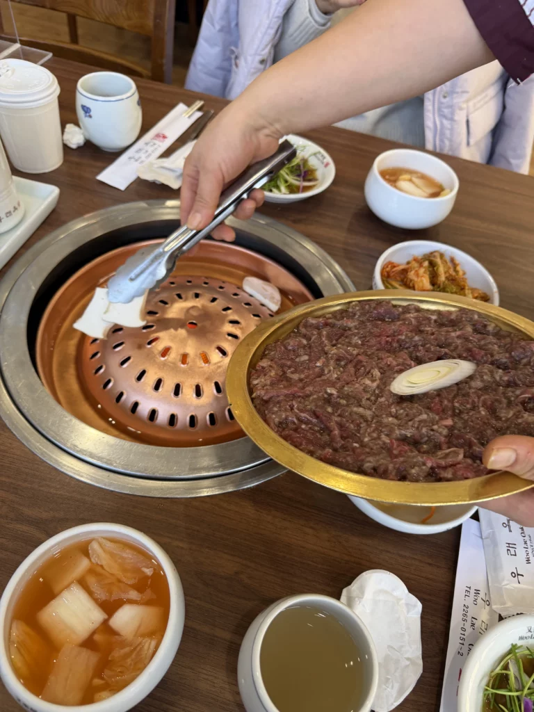 Raw marinated Hanwoo beef bulgogi on a golden serving plate next to traditional copper grill at Woo Lae Oak Seoul, with tongs placing meat for tableside grilling alongside Korean banchan side dishes.