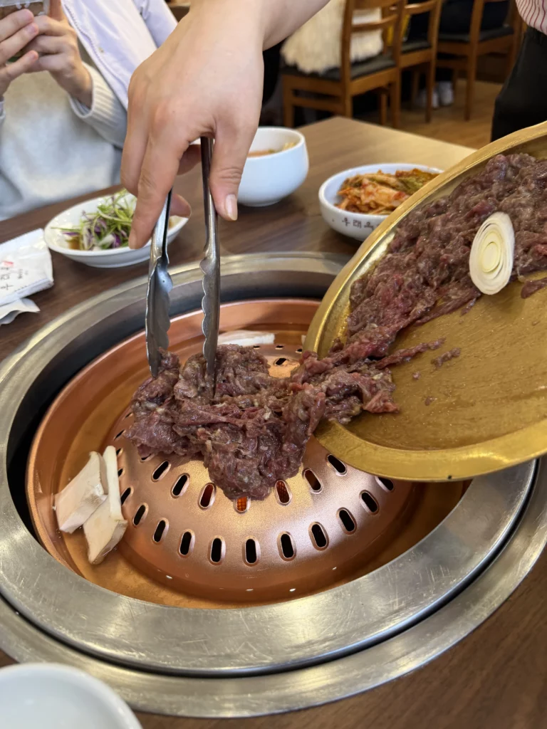 Tongs transferring grilled Hanwoo beef bulgogi from traditional copper grill at Woo Lae Oak Seoul, with raw marinated meat on golden serving plate demonstrating the interactive tableside Korean BBQ cooking experience.