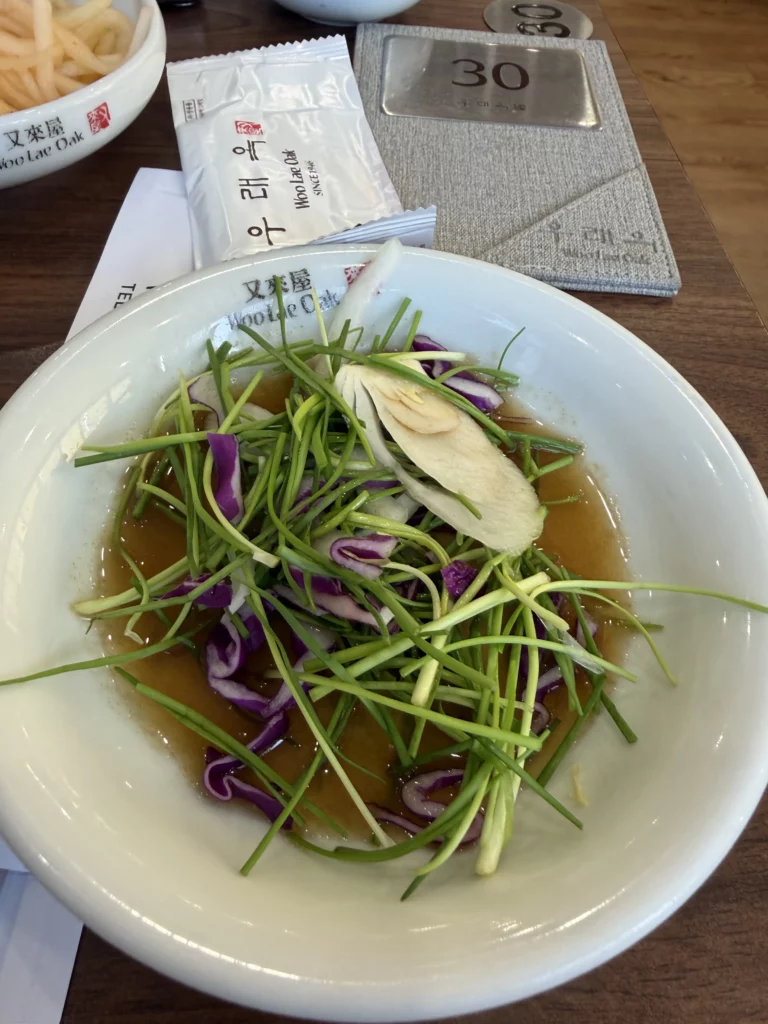 Fresh garnishes for Pyongyang naengmyeon at Woo Lae Oak Seoul, featuring julienned scallions, sliced Korean pear, and purple onions in clear beef broth before adding the traditional buckwheat noodles.